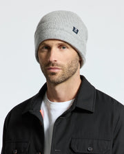 A man is wearing a gray Psycho Bunny MENS RACKED BEANIE - B6A188E200 and a black jacket over a white shirt, standing against a plain background to highlight the simplicity and style of this merino wool beanie.