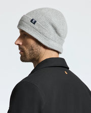 A man sporting a light gray MENS RACKED BEANIE - B6A188E200 by Psycho Bunny and a black jacket faces sideways. The beanie, crafted from warm merino wool, features a small tag on the side. The background is a plain white wall.
