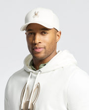 Against a plain light gray background, a person wears the Psycho Bunny MENS NEW CASTLE EMBROIDERED GRAPHIC BASEBALL CAP - B6A547F200 and a white hoodie adorned with bunny logos, exuding an air of quiet luxury.