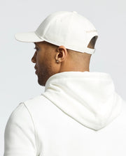 A person is wearing a white hoodie and a Psycho Bunny MENS NEW CASTLE EMBROIDERED GRAPHIC BASEBALL CAP - B6A547F200, with small text on the back. The scene is captured from behind against a plain, light-colored background.