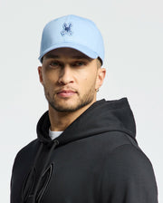 A person wearing a Psycho Bunny MENS NEW CASTLE EMBROIDERED GRAPHIC BASEBALL CAP in light blue, featuring a cartoon logo, faces the camera. They have short facial hair, wear a black hoodie, and stand against a plain white backdrop, embodying understated quiet luxury.