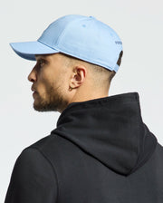 A person in a black hoodie and a Psycho Bunny MENS NEW CASTLE EMBROIDERED GRAPHIC BASEBALL CAP - B6A547F200 in light blue faces left. Featuring subtle side text that conveys quiet luxury, they have a short beard and an earring, standing against a plain, light background.