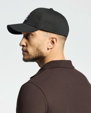 A man wears a Psycho Bunny MENS CLASSIC BASEBALL CAP (B6A816A1HT) in black, perfectly paired with a dark polo shirt. He sports a stud earring in his left ear, gazing sideways against a light background.