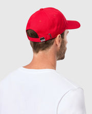 Wearing Psycho Bunny's MENS CLASSIC BASEBALL CAP - B6A816A1HT in red, with its adjustable strap and embroidered bunny logo, pairs perfectly with a white T-shirt. They are turned away from the camera against a gray background, showcasing the cap's six-panel cotton twill.