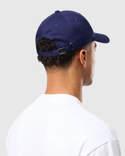 A person in a classic white shirt faces away, wearing the MENS CLASSIC BASEBALL CAP - B6A816A1HT by Psycho Bunny, which is a navy blue six-panel cotton twill cap with an adjustable strap. The background is plain light gray.