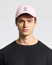 A young person sports Psycho Bunny's MENS CLASSIC BASEBALL CAP - B6A816A1HT made of cotton twill with an embroidered bunny and crossbones logo. Their black shirt features the same playful design, standing out against the plain background.
