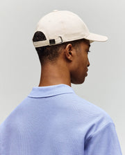 A person wearing the Psycho Bunny MENS CLASSIC BASEBALL CAP - B6A816A1HT in adjustable beige cotton, embroidered with a Bunny, and a light blue collared shirt, shown from behind against a plain light gray background.