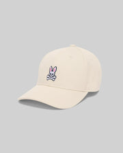 The Psycho Bunny MENS CLASSIC BASEBALL CAP - B6A816A1HT is a beige cotton hat with an embroidered Bunny logo (pink ears above crossed bones) on the front, shown on a plain gray background.