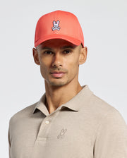 A person wears a Psycho Bunny coral baseball cap with a rabbit and crossbones logo, offering an adjustable fit. They pair it with a classic beige polo shirt featuring the same small rabbit logo. The cotton twill cap contrasts nicely against the neutral background.