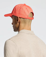 A person in a coral-colored Psycho Bunny MENS CLASSIC BASEBALL CAP - B6A816F200 is viewed from the side and back. Dressed in a beige shirt, they maintain a neutral expression and classic style against a plain, light-colored background.