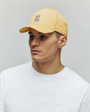 A young man in a Psycho Bunny MENS CLASSIC BASEBALL CAP - B6A816F200 (light yellow, small bunny logo), a white recycled polyester t-shirt, and a small hoop earring looks slightly to the side against a plain background.