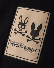 The MENS MONROE TWILL PATCH SCARF - B6A892D200 by Psycho Bunny features a black rectangular patch that displays two bunny skull logos with crossbones underneath. The left bunny skull has a cheerful, open-mouthed expression while the right one has a neutral look. Below the logos, "PSYCHO BUNNY" is written twice in uppercase letters.
