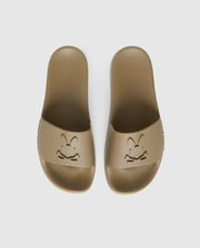 A pair of beige pool slides with a textured sole and upper strap featuring an embossed design. The design consists of a stylized bunny face with crossbones underneath, creating a playful skull and crossbones motif. They come with a molded footbed for added comfort and support, against a plain white background.
Product Name: MENS LOGO SLIDES - B6A910U1FT
Brand Name: Psycho Bunny