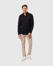 A person is standing against a plain background, wearing a Psycho Bunny MENS ARTHUR JERSEY SHIRT - B6C748D200 made of stretch Pima cotton, beige trousers, and white sneakers. They have short hair and are smiling slightly, with one hand hanging by their side and the other partially in a pocket.