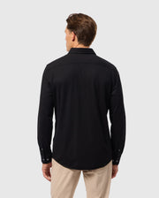 A man with short brown hair is shown from the back, wearing a black long-sleeved Psycho Bunny MENS ARTHUR JERSEY SHIRT - B6C748D200 and beige pants. The background is a plain light gray.