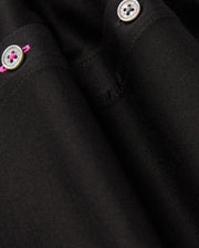 Close-up of the black fabric from Psycho Bunny's MENS ARTHUR JERSEY SHIRT - B6C748D200, featuring two small metallic buttons. The upper buttonhole is accentuated with a pink thread stitch, providing a subtle pop of color against the smooth, finely woven monochromatic textile.