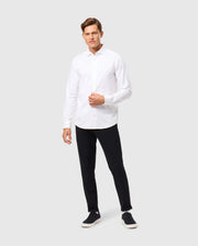 A man wearing a Psycho Bunny MENS ARTHUR JERSEY SHIRT - B6C748D200 in stretch Pima cotton and black pants stands against a plain gray background. He is also wearing black sneakers with white soles, has one hand in his pocket, and is looking at the camera with a neutral expression.