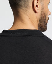 A man with short hair and a beard wears the black Psycho Bunny MENS JOHNNY COLLAR SWEATER, B6E258E200, featuring a subtle embossing near the neckline. This cotton-wool blend piece adds sophistication against a plain, light backdrop.