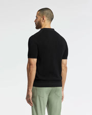 A man with short hair stands facing away, wearing a Psycho Bunny MENS LEETON JOHNNY COLLAR KNIT POLO - B6E562F200 in black and green pants. The 100% cotton shirt showcases an embroidered Bunny on a plain white background that highlights its breathable elegance.