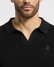A man wears a black 100% cotton ribbed Psycho Bunny MENS LEETON JOHNNY COLLAR KNIT POLO with an embroidered Bunny emblem on the chest. The breathable short-sleeved shirt is set against a plain white background, showing only his torso and part of his face.