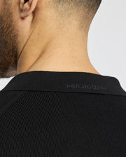A close-up showcases a man's neck and shoulder in a black, breathable 100% cotton MENS LEETON JOHNNY COLLAR KNIT POLO by Psycho Bunny. Its ribbed texture and subtle embroidered Bunny logo near the collar enhance its design.