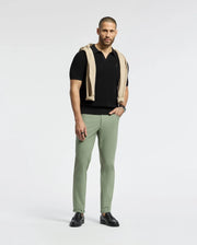 A man stands against a plain white background in the breathable black MENS LEETON JOHNNY COLLAR KNIT POLO by Psycho Bunny, sporting sage green pants and black shoes. A beige sweater with an embroidered bunny accent is casually draped over his shoulders. His hair is neatly cut, with a trimmed beard.