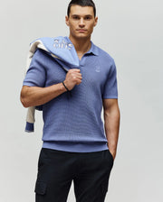 A man wears the Psycho Bunny MENS LEETON JOHNNY COLLAR KNIT POLO (B6E562F200) in light blue with dark pants, standing against a plain background, one hand in his pocket and a light blue and white jacket over his shoulder.