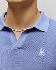 A person wears a Psycho Bunny MENS LEETON JOHNNY COLLAR KNIT POLO (B6E562F200) in light blue, featuring a textured finish and bunny logo, paired with a thin silver chain necklace; only their lower face and upper torso are visible.