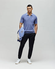 A man stands against a plain background in a light blue Psycho Bunny MENS LEETON JOHNNY COLLAR KNIT POLO (B6E562F200), black pants, and white sneakers, holding a blue and white jacket over his left arm as he looks slightly right.