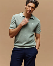 A man in a slim fit light green Psycho Bunny MENS LEETON JOHNNY COLLAR KNIT POLO with an embroidered chest Bunny and dark pants stands against a beige background, looking to the side with one hand touching his collar.