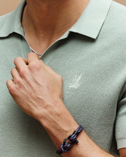 A person wears the Psycho Bunny Mens Leeton Johnny Collar Knit Polo (B6E562F200) in light green, featuring an embroidered chest Bunny, paired with a silver chain necklace and blue-and-black knotted bracelet while holding the necklace.