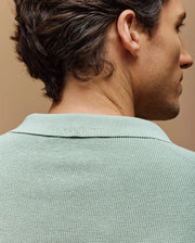 A man is seen from behind in a slim fit light green MENS LEETON JOHNNY COLLAR KNIT POLO by Psycho Bunny, featuring subtle logo embroidery on the collar against a warm, neutral background.