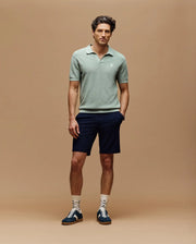 A man with short, styled hair stands against a beige background, wearing the Psycho Bunny MENS LEETON JOHNNY COLLAR KNIT POLO in light green, navy shorts, white patterned socks, and navy sneakers with white laces. His hands are in his pockets.