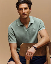 A man with wavy brown hair wears the Psycho Bunny MENS LEETON JOHNNY COLLAR KNIT POLO (B6E562F200) in light green and a bracelet, sitting on a wooden chair against a beige background, looking to the side and smiling softly.
