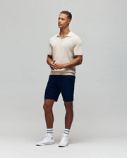 Against a plain backdrop, a person wears the Psycho Bunny MENS LEETON JOHNNY COLLAR KNIT POLO in beige, paired with dark shorts, white high-top sneakers, and white socks featuring black stripes. They stand gazing sideways with hands loosely clasped.