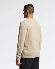 A man with short dark hair is facing away, wearing a beige MENS LINCOLN MERINO WOOL SWEATER (B6E831E200) by Psycho Bunny and black pants. The background is plain white.