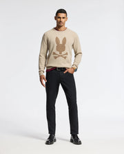 A man stands against a plain background wearing the Psycho Bunny MENS LINCOLN MERINO WOOL SWEATER (B6E831E200) in beige, featuring a large graphic of a rabbit over crossbones. He pairs it with black pants and black shoes. He has short hair and is looking at the camera with a neutral expression.