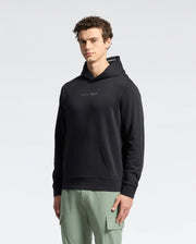 A man with short curly hair wears a black Psycho Bunny Mens Nora Classic French Terry Wordmark Hoodie with an embroidered chest logo and light green cargo pants, standing against a plain light background.