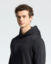A young man with short, curly brown hair wears a Psycho Bunny MENS NORA CLASSIC FRENCH TERRY WORDMARK HOODIE (B6H434F200) in black, looking toward the camera against a plain light background.