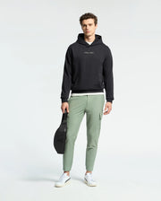 A man faces forward against a plain light background, wearing a black Psycho Bunny MENS NORA CLASSIC FRENCH TERRY WORDMARK HOODIE, light green joggers, white sneakers, and holding a black bag in his left hand.