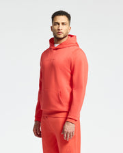 A man with short hair wears matching coral French terry Psycho Bunny hoodie and sweatpants, featuring subtle wordmark embroidery. He stands against a plain white background, gazing at the camera with a neutral expression.