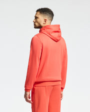 A person stands sideways in a bright red Psycho Bunny MENS NORA CLASSIC FRENCH TERRY WORDMARK HOODIE (B6H434F200) with matching pants against a plain white background. The hood is down, showcasing subtle embroidery, with their hands by their sides.