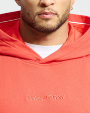 A person wears a coral MENS NORA CLASSIC FRENCH TERRY WORDMARK HOODIE by Psycho Bunny, with "Psycho Bunny" embroidery. The hood reveals a white lining, and their partially visible face shows a beard and earring.
