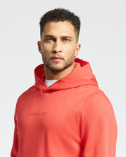 A man stands confidently against a white background, wearing the Men's Nora Classic French Terry Wordmark Hoodie - B6H434F200 by Psycho Bunny. The vibrant orange hoodie with "Psycho Bunny" embroidered boldly across the chest complements his short, curly hair and beard.