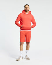 A man wears a matching coral ensemble, featuring the Psycho Bunny Nora Classic French Terry Wordmark Hoodie and shorts. His look is completed with white socks and beige sneakers against a plain off-white background.