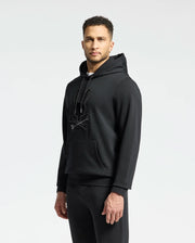 A man confidently stands against a plain background wearing the MENS NEW CASTLE EMBROIDERED FRENCH TERRY HOODIE by Psycho Bunny. The black hoodie pairs seamlessly with his pants, featuring a stylized bunny logo and front pocket. He sports short, curly hair and a trimmed beard.