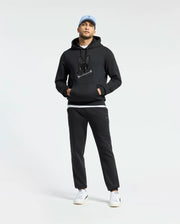 A man stands against a plain white background wearing a Psycho Bunny MENS NEW CASTLE EMBROIDERED FRENCH TERRY HOODIE in black, black joggers, a light blue cap, and white sneakers.