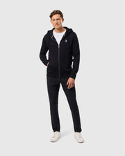 A person wearing a stylish Psycho Bunny MENS LEVY ZIP FLEECE HOODIE - B6H720D200 in black over a white T-shirt, paired with black pants and crisp white sneakers, stands against a plain light grey background.