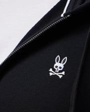 Close-up of the Psycho Bunny MENS LEVY ZIP FLEECE HOODIE - B6H720D200 in black, featuring a sleek white zipper and an embroidered cartoon rabbit skull and crossbones design on the fabric.