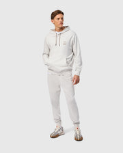 A person wearing a Psycho Bunny MENS JAMES PREMIUM HOODIE - B6H789D200 in light gray, along with matching sweatpants, is standing against a plain background. The hoodie features an embroidered bunny on the chest. The ensemble is complemented by gray and white sneakers, one hand resting casually in a pocket as the individual exudes a relaxed demeanor.